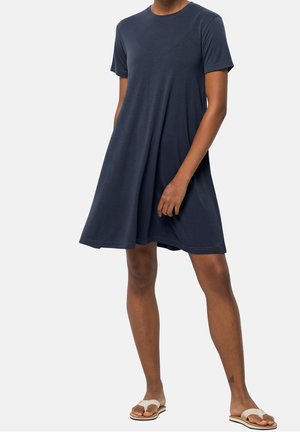 Žena v tmavě modrých šatech s krátkými rukávy po kolena a béžových sandálech, stojící s jednou rukou u boků.