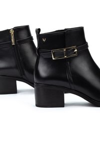 Botas de tobillo de cuero negro con tacón cuadrado, detalle de hebilla dorada y cremallera lateral. Textura suave y diseño moderno.