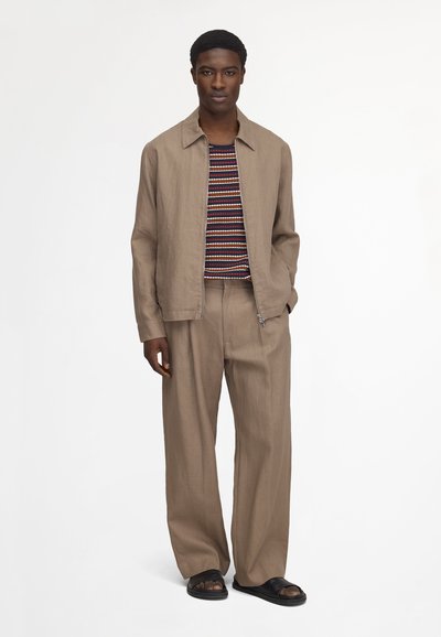 Homme debout portant une veste beige à fermeture éclair, un pantalon large, une chemise rayée et des sandales noires sur un fond blanc uni.