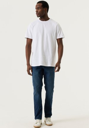 Mann trägt ein schlichtes weißes kurzärmliges T-Shirt, blaue Jeans und weiße Turnschuhe, steht und schaut nach links.