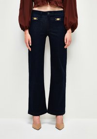Jeans de denim oscilo con pinzas oscura y detalles en dorado en la cintura, que presentan un diseño clásico de cinco bolsillos y una sutil textura.