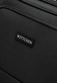Černý texturovaný kufr s viditelným zipem a obdélníkovou černou etiketou, na které je bílými písmeny napsáno jméno značky "WITTCHEN".