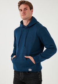 Marineblaue Kapuzenpullover aus weichem Material, mit einer Fronttasche, verstellbarem Kordelzug an der Kapuze und einem kleinen Logo-Tag nahe dem Saum.