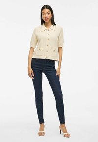 Beige gebreid vest met korte mouwen en een kraag, met knopen aan de voorkant en twee zakken, gecombineerd met donkere skinny jeans en lichte sandalen.