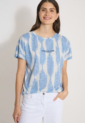 Femme aux longs cheveux foncés porte une chemise à motifs bleu clair et blanc avec le texte "SAND AND TIME", associée à un pantalon blanc, main dans la poche.