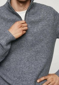 Pull gris à fermeture éclair avec une texture côtelée, un col montant et des manches longues. Le tissu semble doux et ajusté, présentant un design simple.