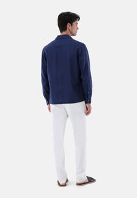 Camicia di lino blu marino con maniche lunghe, dettagli con bottoni e taglio dritto, abbinata a pantaloni bianchi e sandali neri.