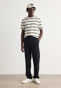 Stribet beige t-shirt med marineblå vandrette striber, sorte bukser og hvide sneakers. En cremefarvet kasket med et logo fuldender looket.