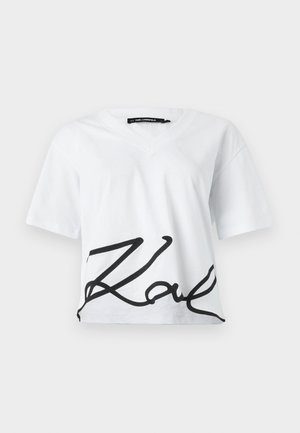 Camiseta blanca recortada con escote en V y mangas cortas. Presenta un gran logotipo cursivo negro en la parte inferior delantera. Está hecha de algodón.