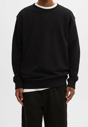 Mann trägt einen schlichten schwarzen Langarm-Sweatshirt über einem weißen T-Shirt mit schwarzen Hosen, steht vor weißem Hintergrund.