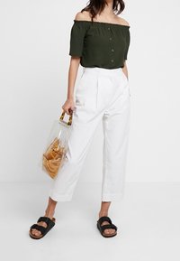 Tummanvihreä olkaimeton paita napilla, valkoiset leveät housut, mustat soljilla varustetut sandaalit ja läpinäkyvä käsilaukku.