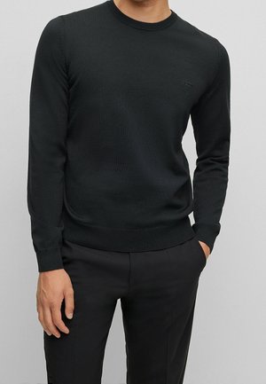 Mand iført en tætsiddende sort langærmet crewneck-sweater med subtilt "BOSS"-logo på brystet, kombineret med sorte bukser.