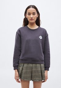 Bluza mov are un decolteu rotund și manșete drepte, decorată cu un grafic de buze gri pe piept. Este asortată cu o fustă plisată în carouri.