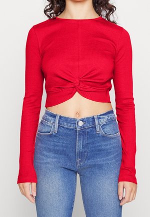 Kobieta w długim rękawie, nosząca czerwoną krótką bluzkę z skręconym węzłem z przodu oraz niebieskie dżinsy z wysokim stanem, stojąca na tle jednolitym.