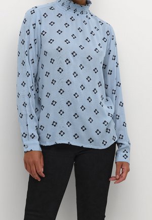 Light blue long-sleeve blouse with black floral diamond pattern and ruffled high neckline, paired with black pants on a standing person.