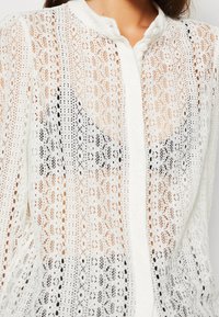 Vit spetsblus med knäppning framtill, med intrikata blommönster och geometriska mönster, hög krage och semi-genomskinlig textur.