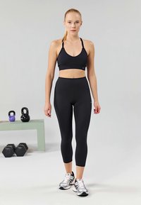 Crna sportska grudnjak i tajice od rastezljivog materijala, koje imaju prilagođen dizajn. Model nosi bijele i sive atletsku obuću. Fitness oprema u pozadini.