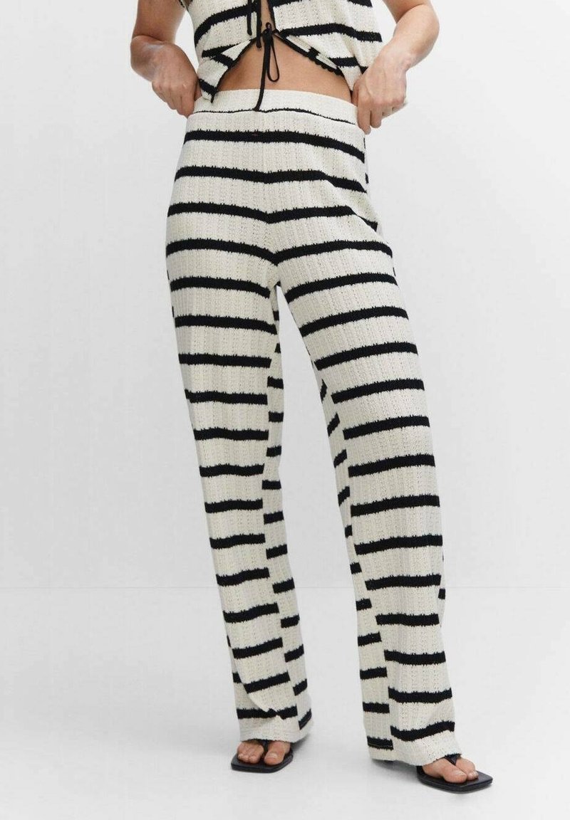 Personne portant un pantalon en maille blanc taille haute avec des rayures horizontales noires irrégulières et des sandales noires à bout carré sur un fond uni.