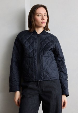 Kvinde iført en sort quiltet jakke med lynlås og mørke bukser, stående mod en lysegrå væg og kigger let mod venstre.