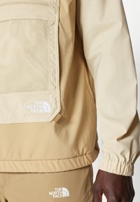 Chaqueta beige con bolsillo de malla, puños elásticos y parche con logo. Textura suave, tejido ligero y diseño deportivo.