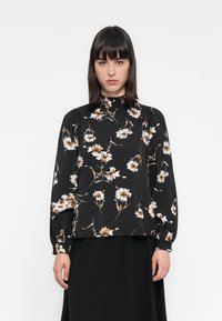 Svart bluse med lange ermer og blomstermønster i krem og beige, med høy hals og rynkede detaljer på skuldrene og mansjettene.