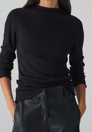 Persoon draagt een zwart shirt met lange mouwen en gekartelde randen, met zwarte hooggetailleerde leren broek en handen in de zakken.