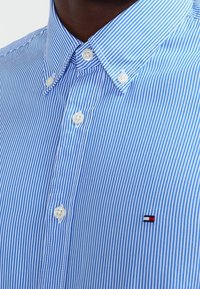 Camisa de cuello con rayas azules y blancas, con cierre de botones, que presenta botones redondos blancos y un pequeño logo en el lado izquierdo del pecho.