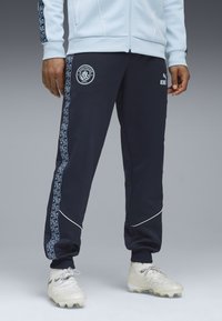 Marineblaue Jogginghose mit einem blauen Muster an der Seite, versehen mit einem Manchester City-Logo und weißen Paspeln. Hergestellt aus weichem, flexiblem Material.
