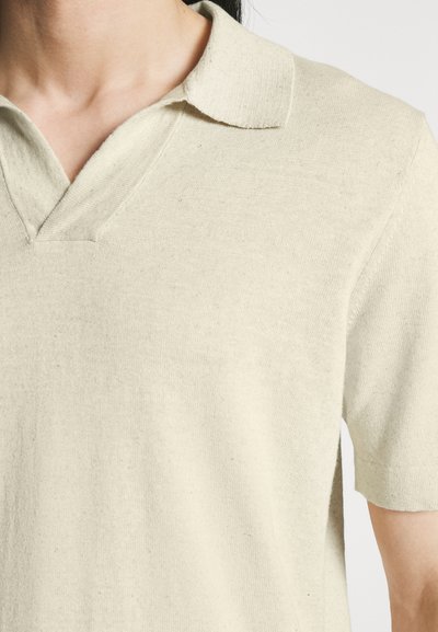Personne portant un polo en maille beige à manches courtes avec un col en V et un col doux.