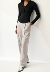 Maglietta henley nera a maniche lunghe con chiusura a bottoni, abbinata a pantaloni plissettati grigio chiaro e sneakers in bianco e nero, su uno sfondo neutro.