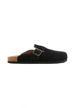 Sort slip-on træsko med justerbar rem og sølvspænde, korksål og flad sål, vist fra siden på hvid baggrund.