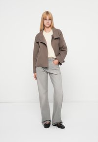 Veste légère marron avec col replié, associée à un pull crème et un jean gris à jambes larges. Le modèle se tient debout, les mains dans les poches.