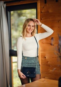 Bílý dlouhý rukávový top, zelená kostkovaná mini sukně se zlatými knoflíky a malá černá crossbody taška s kovovým akcentem.