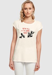 Cream-colored cotton t-shirt with short rolled sleeves featuring a graphic design of people and the text "WE'RE ALL IN THE SAME BOAT" in red.