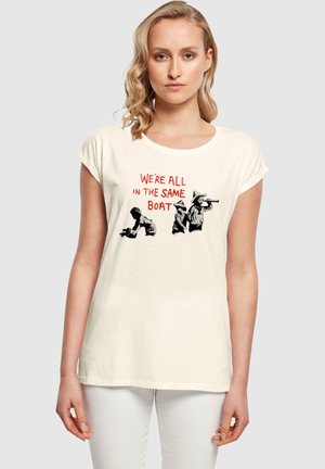 Cremefarbenes Baumwoll-T-Shirt mit kurzen, hochgerollten Ärmeln, das ein grafisches Design von Menschen und den Text "WIR SIND ALLE IM GLEICHENBOOT" in Rot zeigt.