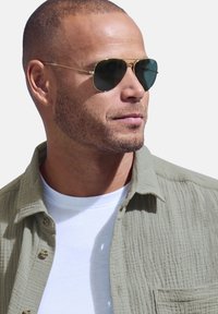 Occhiali da sole rotondi con lenti verdi e montatura in metallo dorato. Indossati con una camicia di un verde oliva testurizzato sopra una t-shirt bianca.