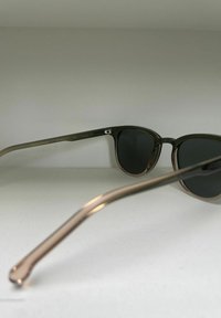 Lunettes de soleil marron et vert avec des verres foncés. Branches en métal avec une légère courbure, portant des gravures de marque et des indications CE.