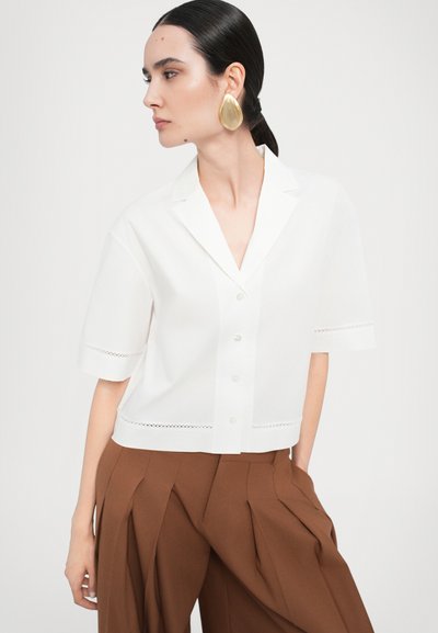 White short-sleeve button-up shirt with a notched collar, featuring eyelet detailing on sleeves. Model wears brown pleated trousers.