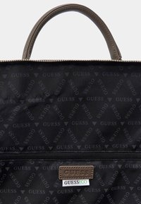 Schwarze Nylon-Tascheninnenseite mit wiederholtem "GUESS"-Muster, braunem Lederetikett, Reißverschluss und doppelt genähten Griffen.