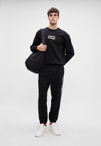 Juodas džemperis su "calvin klein" logotipu, derinamas su juodomis kelnėmis ir baltais sportbačiais su raudonais akcentais, nešamas su juoda rankine.