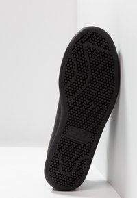 Svart sula för sneakers med ett texturerat gummimönster och en logotyp vid hälen, designad för grepp och fäste på olika ytor.