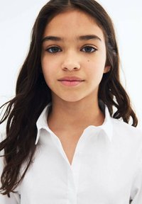 Valkoinen kauluspaita, jossa on pitkät hihat, sileä pinta ja rento istuvuus; päällä lapsella, jolla on pitkät, tummat hiukset ja neutraali ilme.