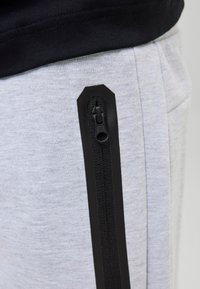 Nahaufnahme eines schwarzen vertikalen Reißverschlusses an hellgrauen Jogginghosen, getragen zu einem schwarzen Oberteil.