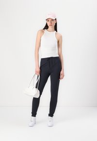 Hvit ribbestrikket tanktop, marineblå joggebukser, hvite joggesko og en rosa caps. Modellen holder en liten hvit håndveske med buede designdetaljer.