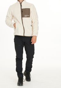 Chaqueta de forro polar en blanco roto con un bolsillo en el pecho marrón y cierre de cremallera, combinada con pantalones negros y botas de senderismo negras. Tejido texturizado.
