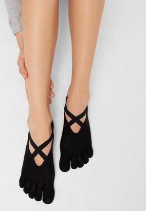 Beine mit schwarzen Zehensocken und gekreuzten elastischen Bändern, eine Hand berührt sanft die linke Wade vor schlichtem Hintergrund.