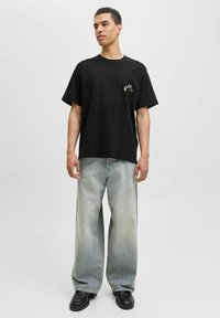 Sort bomulds T-shirt med et lille guld-design, parret med slidte, vide lyseblå jeans og sorte sko med kraftig sål.