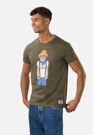 Ung mand iført olivengrøn t-shirt med tegneseriehund iført hipster-tøj, kombineret med blå jeans.