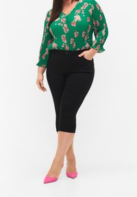 Blusa verde floreale a maniche lunghe con una texture a coste, abbinata a pantaloni neri a tre quarti. L'outfit presenta un paio di tacchi a punta rosa.