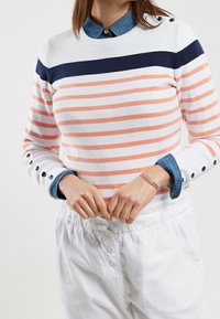 Gestreifter Pullover in Weiß, Koralle und Marineblau, mit Rundhalsausschnitt, Knopfdetails an den Ärmeln und kombiniert mit weißen Hosen.